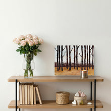 A horizontal canvas art print depicting a forest of tall black tree trunks against a warm brown and gold ground, styled on a wooden shelf next to a vase of pink roses and a candle.