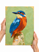 A square canvas painting of a kingfisher perched on a tree stump, held up by two hands against a plain white background. The painting features textured impasto brushstrokes in vibrant blue, orange, green, and gold tones.