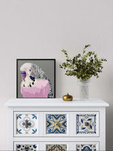 A framed art print of a pink bird styled on a white cabinet with decorative tiles, next to a small gold object and a vase of green leaves, against a light gray wall.